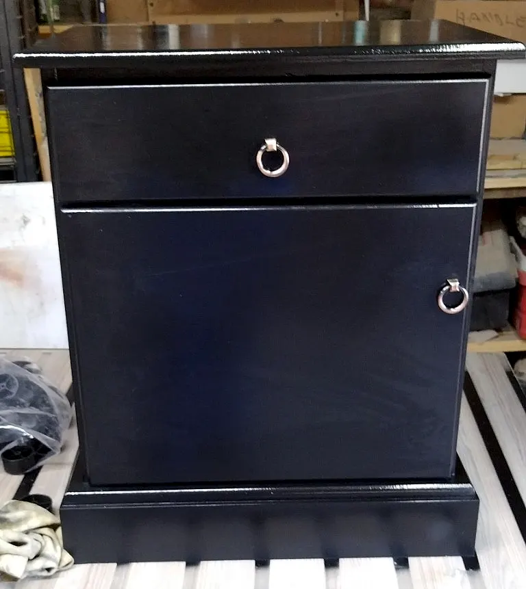 Black bedside cabinet with one drawer and one door with ring handles, standing on a plinth