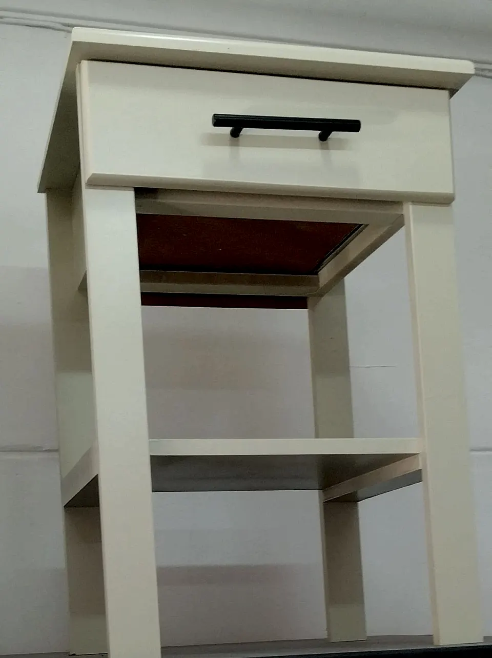 Cream bedside table with one drawer, a black bar handle, a lower shelf and straight legs